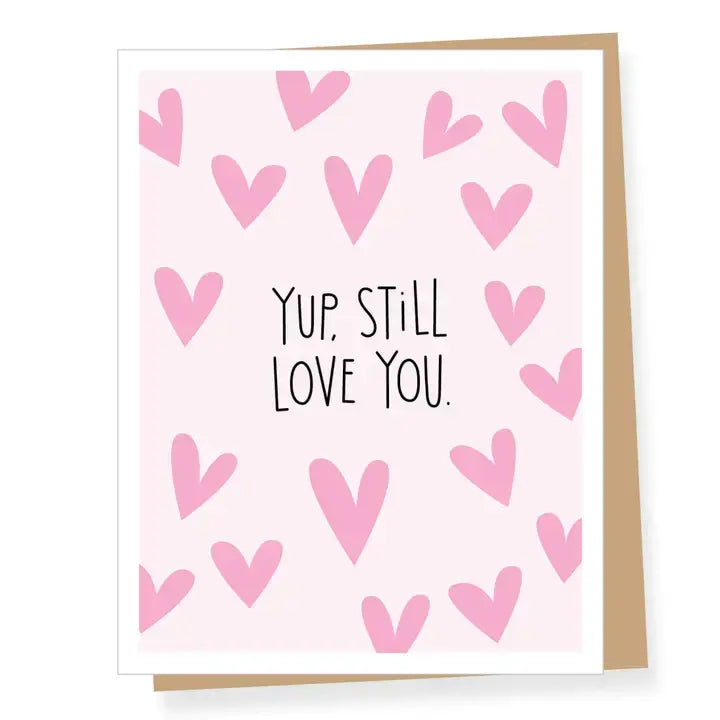 A square greeting card with a light pink background, decorated with scattered pink hearts and the handwritten text 'YUP, STILL LOVE YOU.' in black.
