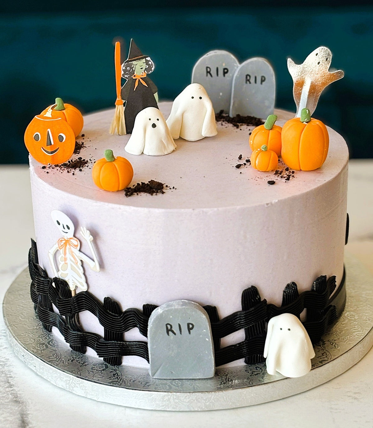 A round Halloween cake with pale pink frosting, decorated with orange pumpkins, white ghosts, a black fence, tombstones, a witch figure, and cookie crumbs, shown on a silver cake board.