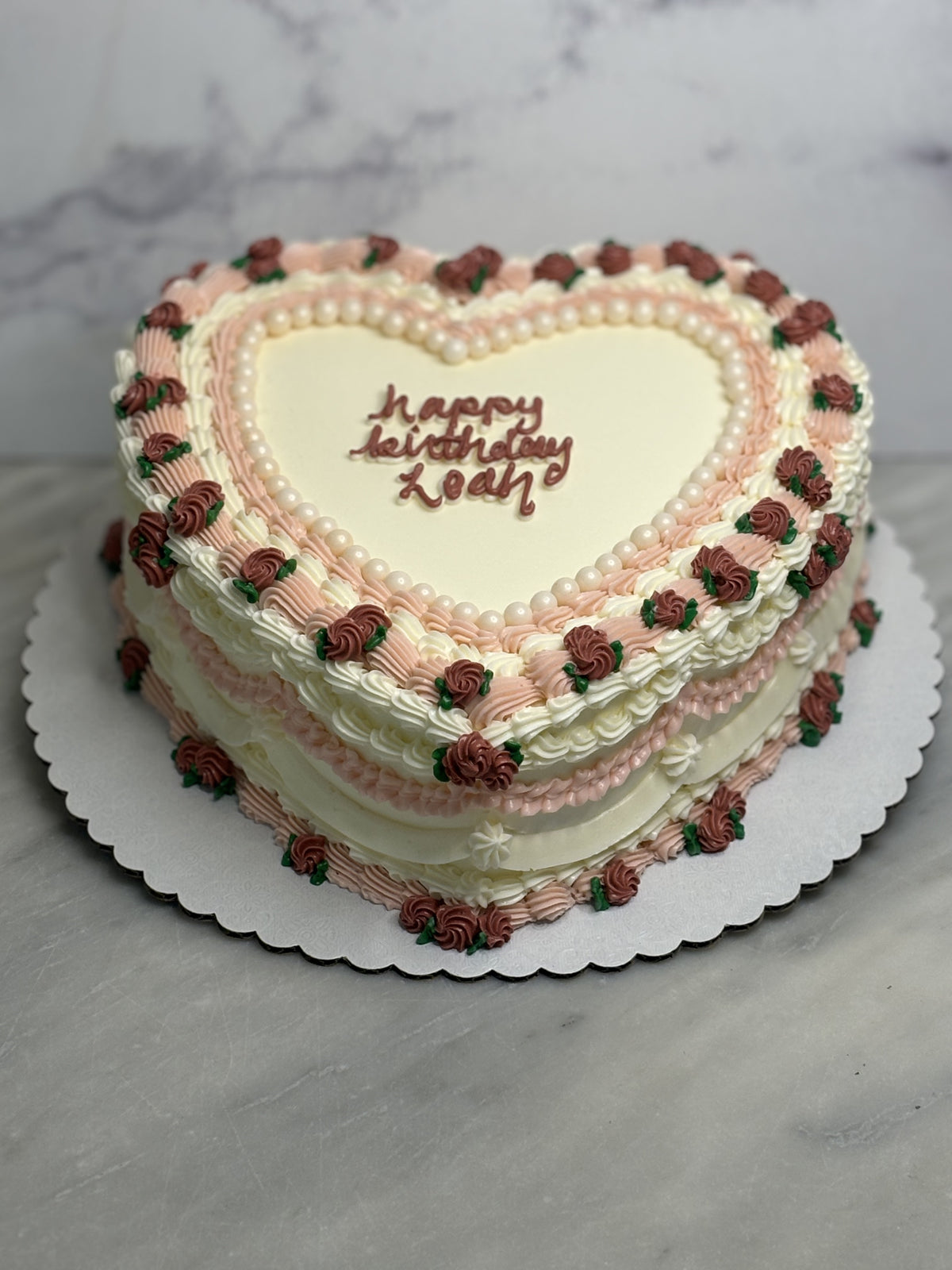 A heart-shaped birthday cake with white and pink frosting, decorated with piped rose motifs in brown and green, and 'Happy Birthday Zoa!' written in brown icing on the top, displayed on a white scalloped cake board.