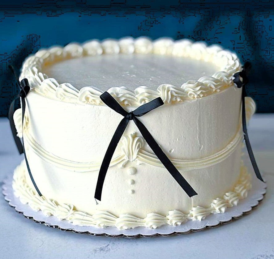 A round white frosted cake with piped decorative borders and black ribbon bows, displayed on a white board against a dark blue background.