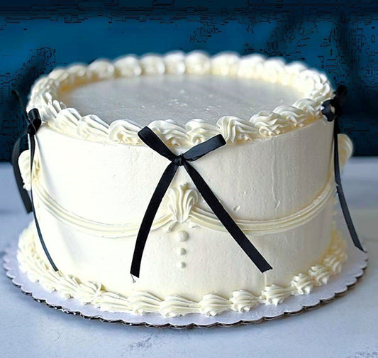 A round white frosted cake with piped decorative borders and black ribbon bows, displayed on a white board against a dark blue background.