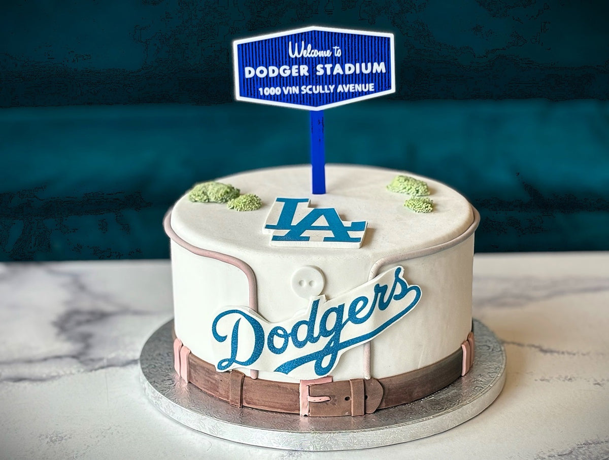 A round white cake decorated to look like a vintage Dodgers baseball jacket, with blue script 'Dodgers' lettering, a blue 'LA' logo, a brown belt detail, and a small sign topper reading 'Welcome to DODGER STADIUM'.