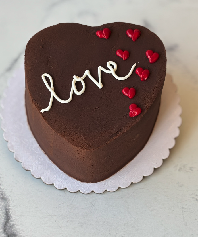 A heart-shaped chocolate cake decorated with white piped icing spelling 'love' and small red heart candies, displayed on a white scalloped cake board.