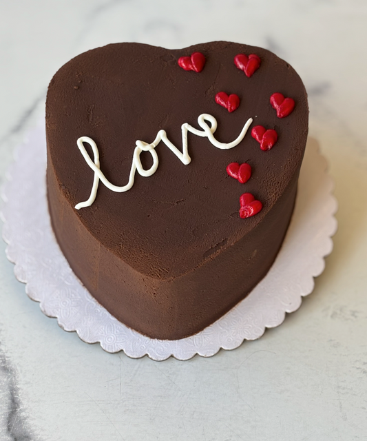 A heart-shaped chocolate cake decorated with white piped icing spelling 'love' and small red heart candies, displayed on a white scalloped cake board.