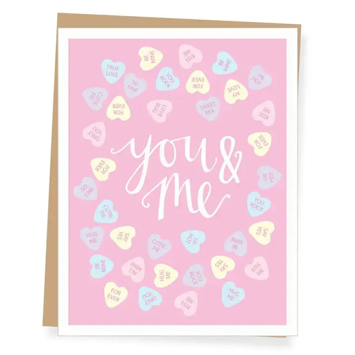 A pink greeting card with white script text reading 'you & me' surrounded by pastel candy hearts, shown with a kraft envelope behind it on a white background.