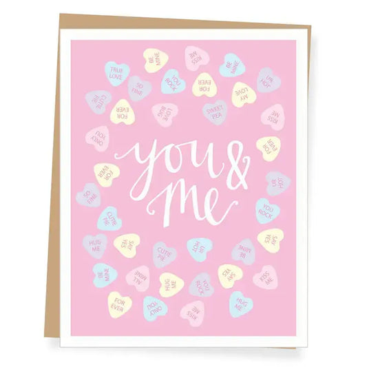 A pink greeting card with white script text reading 'you & me' surrounded by pastel candy hearts, shown with a kraft envelope behind it on a white background.