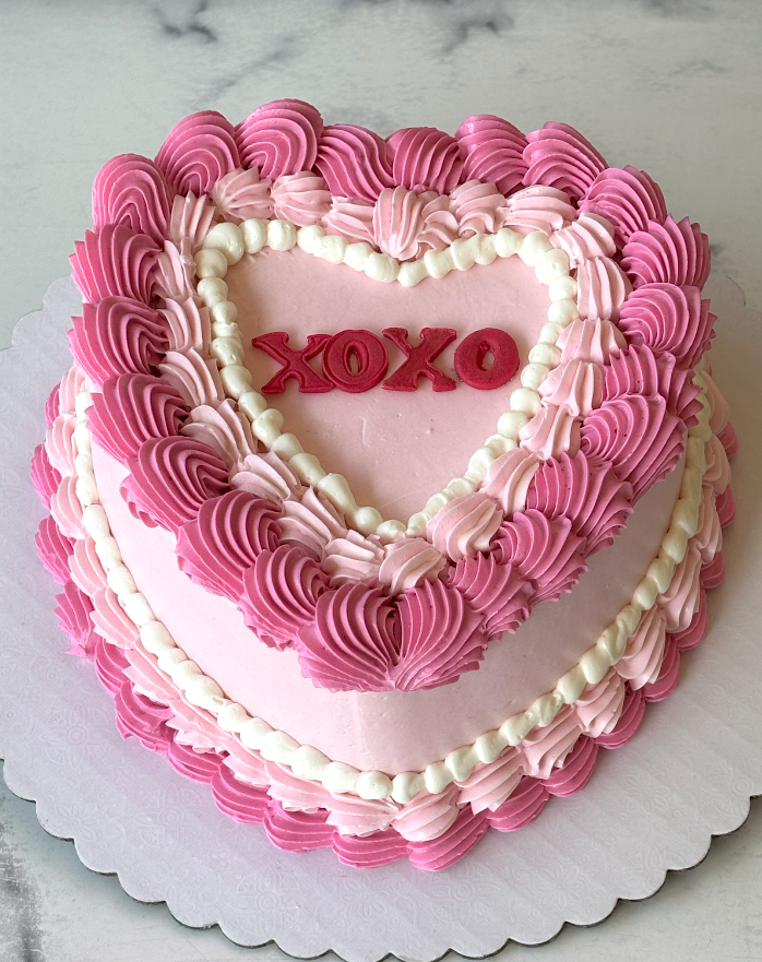 A heart-shaped frosted cake with pink and white piped borders, red 'XOXO' lettering in the center, and a scalloped white cake board on a light marble surface.