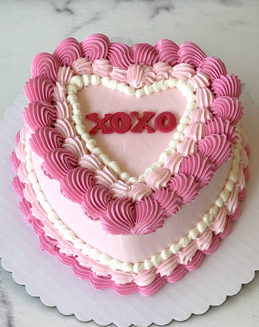 A heart-shaped frosted cake with pink and white piped borders, red 'XOXO' lettering in the center, and a scalloped white cake board on a light marble surface.