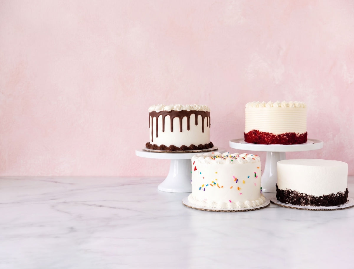 Four decorated cakes displayed on white cake stands against a soft pink wall and light marble surface.